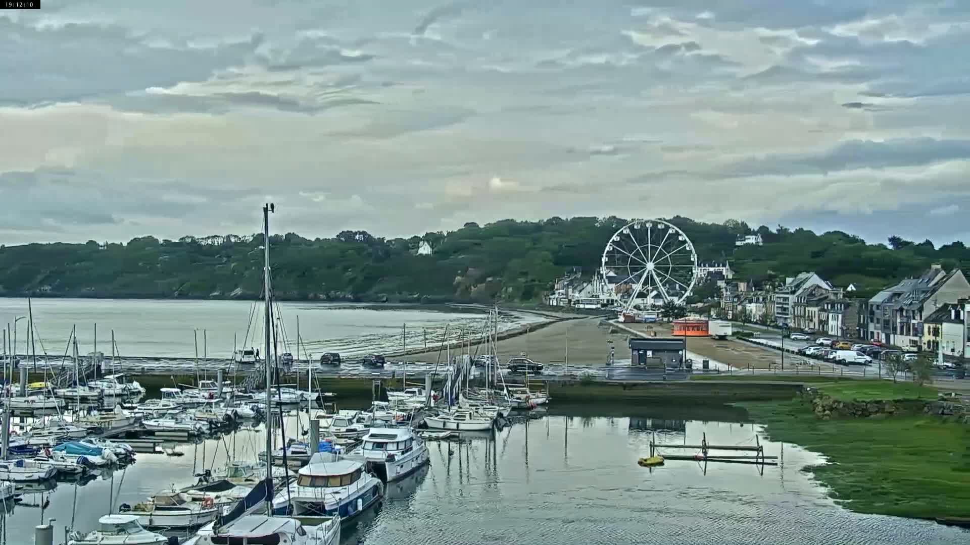 A marina filled with sailboats and motorboats is situated near a town with a Ferris wheel, under a cloudy sky.