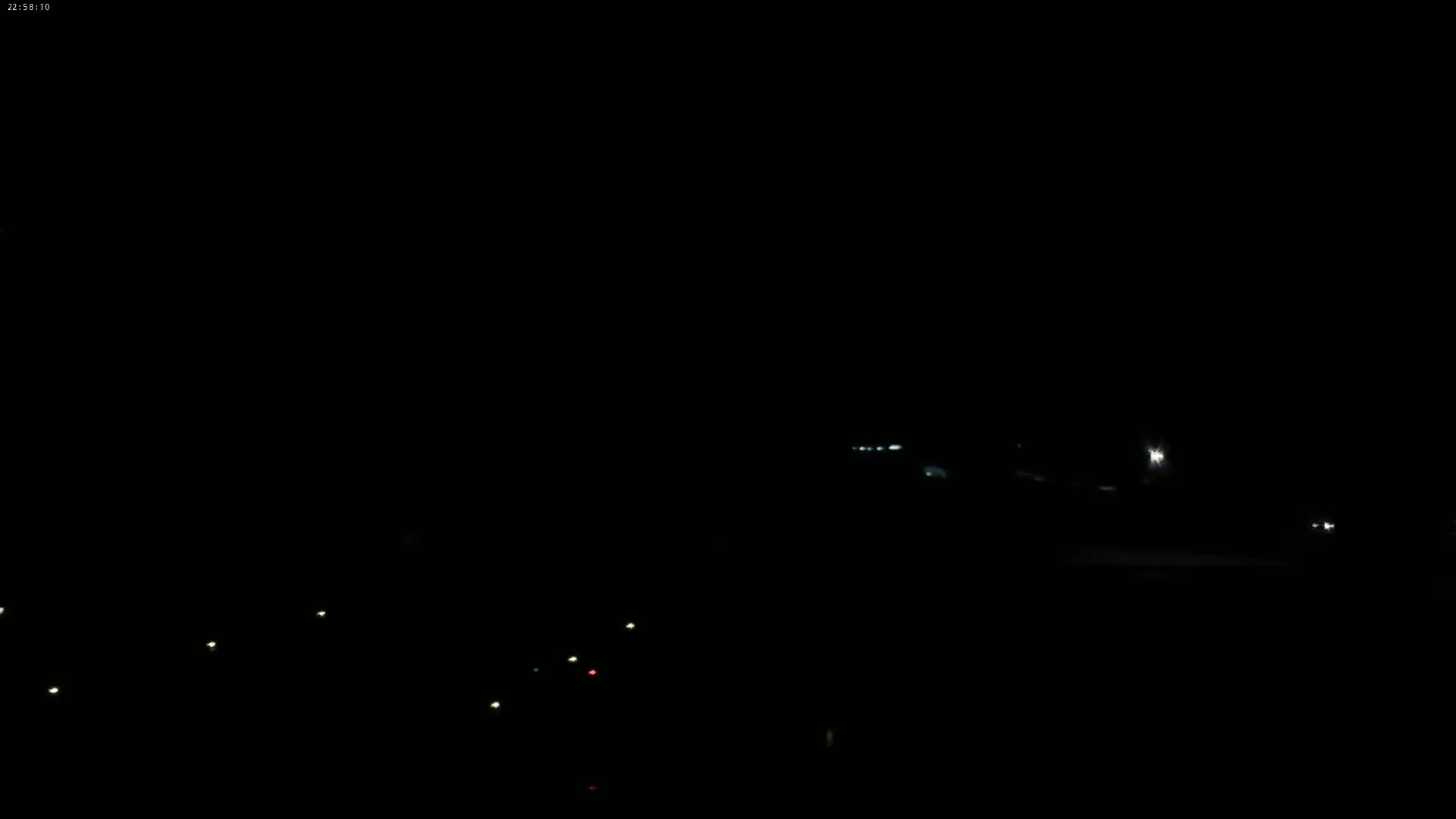 A nighttime, dark scene shows a few distant lights on a road and a scattering of closer, faint lights in the lower part of the frame.