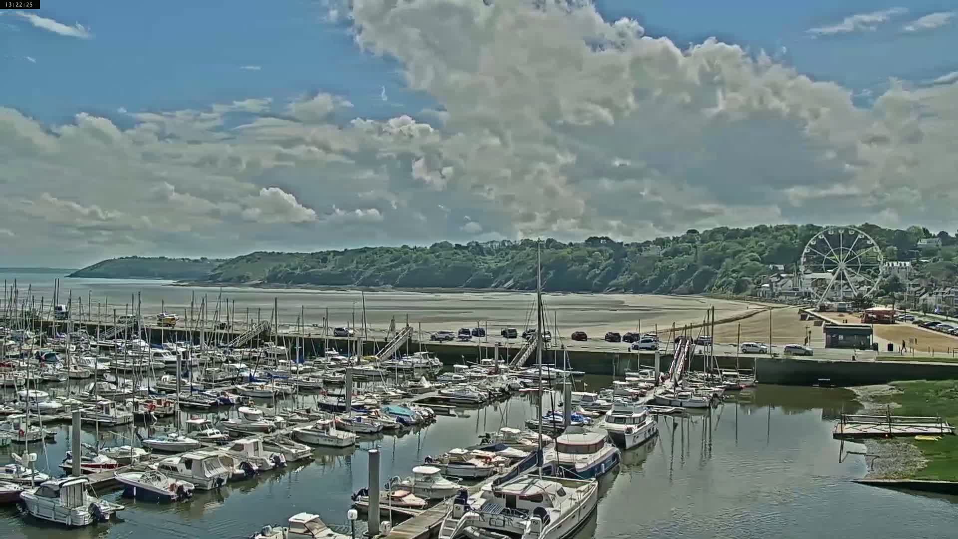 A partly cloudy day reveals a marina full of boats, a sandy beach, a Ferris wheel, and green hills in the background.
