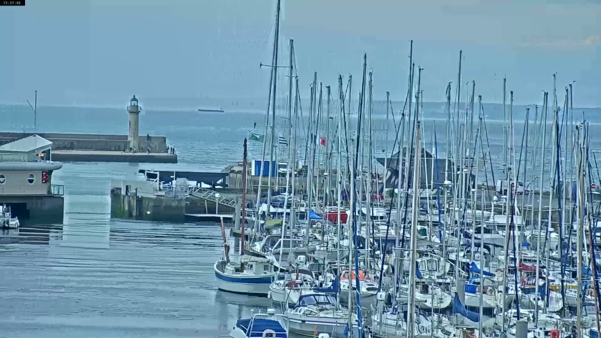 Many sailboats are docked in a harbor on a cloudy day, with a lighthouse visible in the distance.