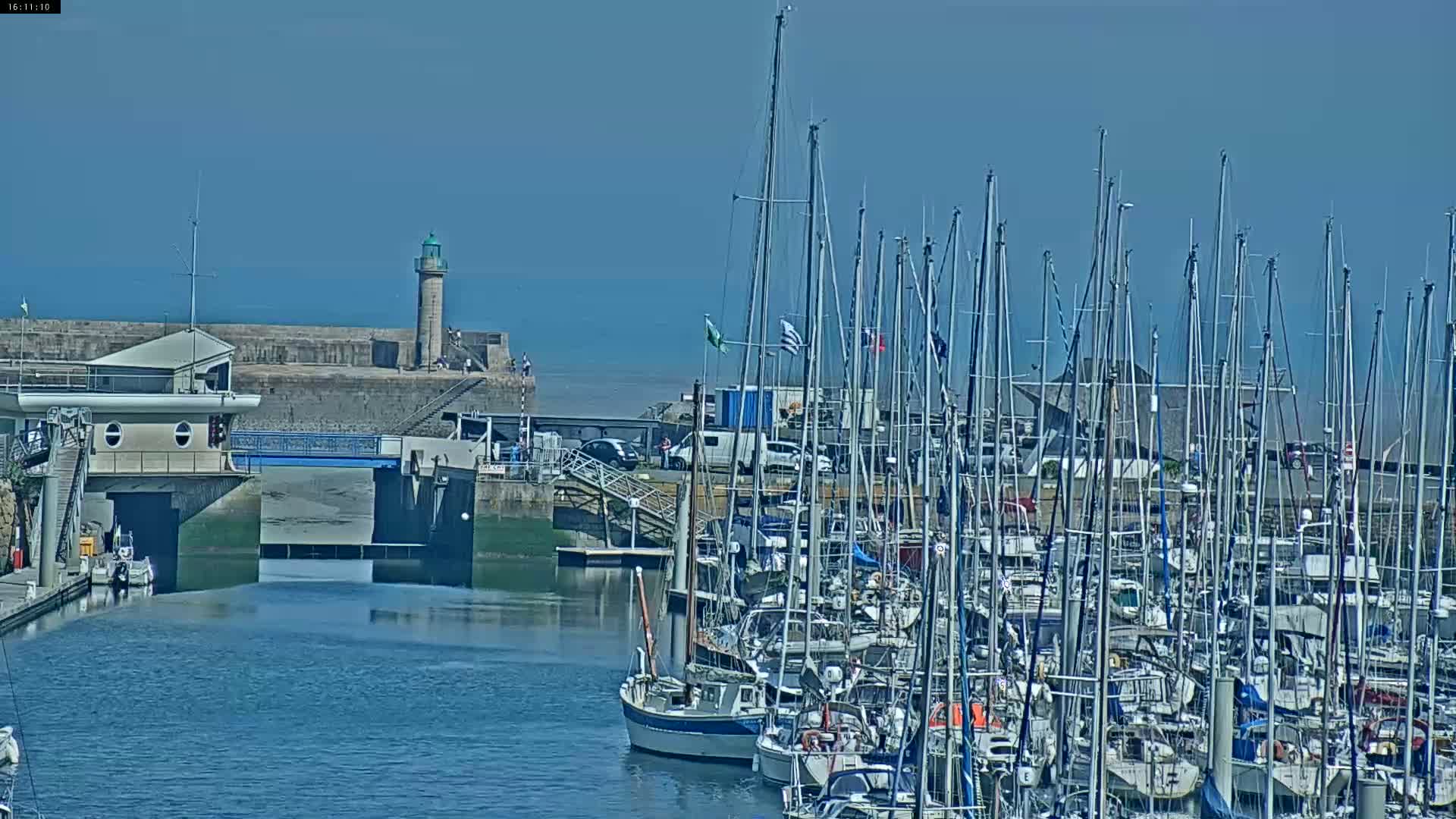 A marina filled with numerous sailboats is seen on a sunny day, with a lighthouse and stone seawall visible in the background.