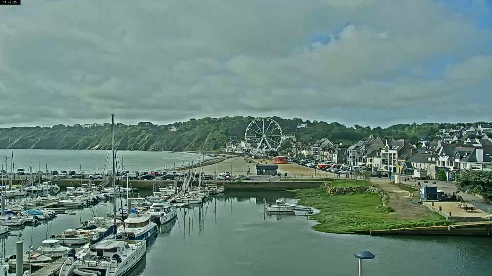 A marina filled with sailboats and motorboats is situated next to a beach and a town with a Ferris wheel under a cloudy sky.
