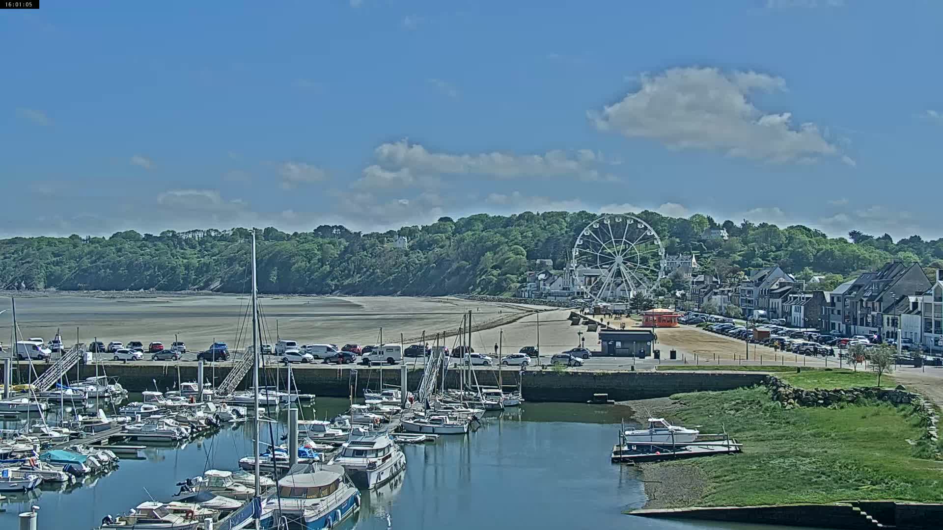 A marina filled with boats sits beside a sandy beach and a town with a Ferris wheel under a partly cloudy sky.