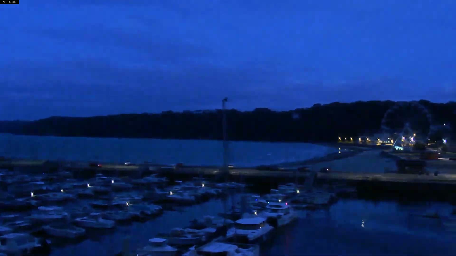 Under a dark blue twilight sky, a marina filled with boats sits before a calm body of water and a dark, tree-covered shoreline.