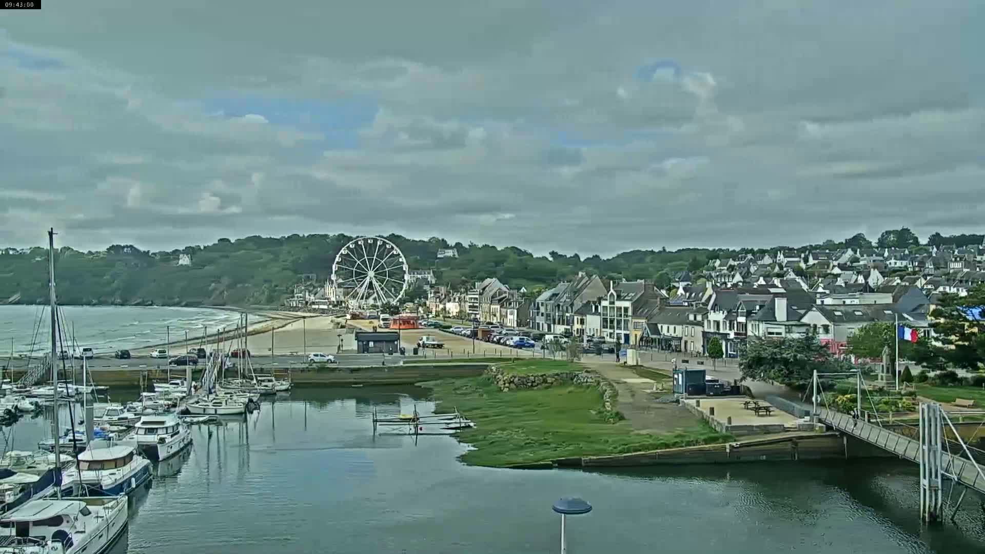 A harbor filled with boats is situated near a town with a Ferris wheel, under a cloudy sky and with a gently wavy ocean visible in the background.