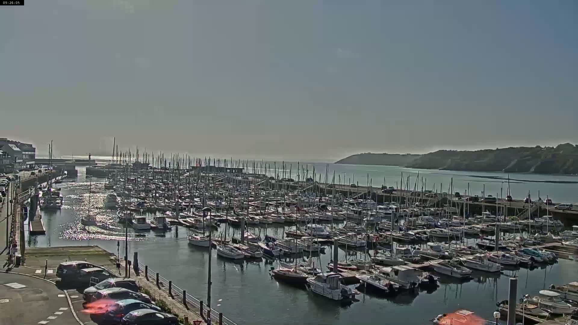 A large marina filled with numerous sailboats and motorboats is situated on calm water under a bright, sunny sky.