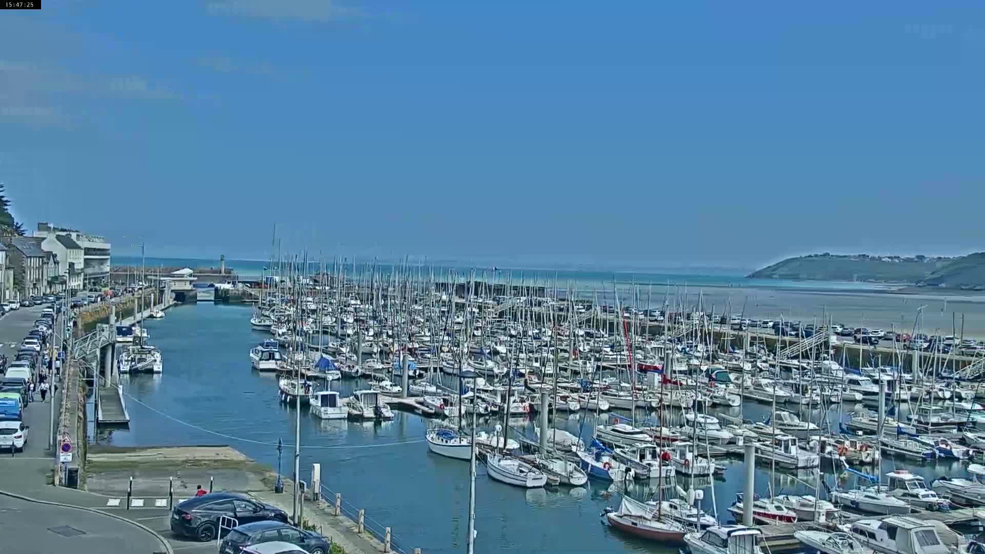 A harbor filled with numerous sailboats and motorboats is shown under sunny skies, with a town and sandy beach visible in the background.