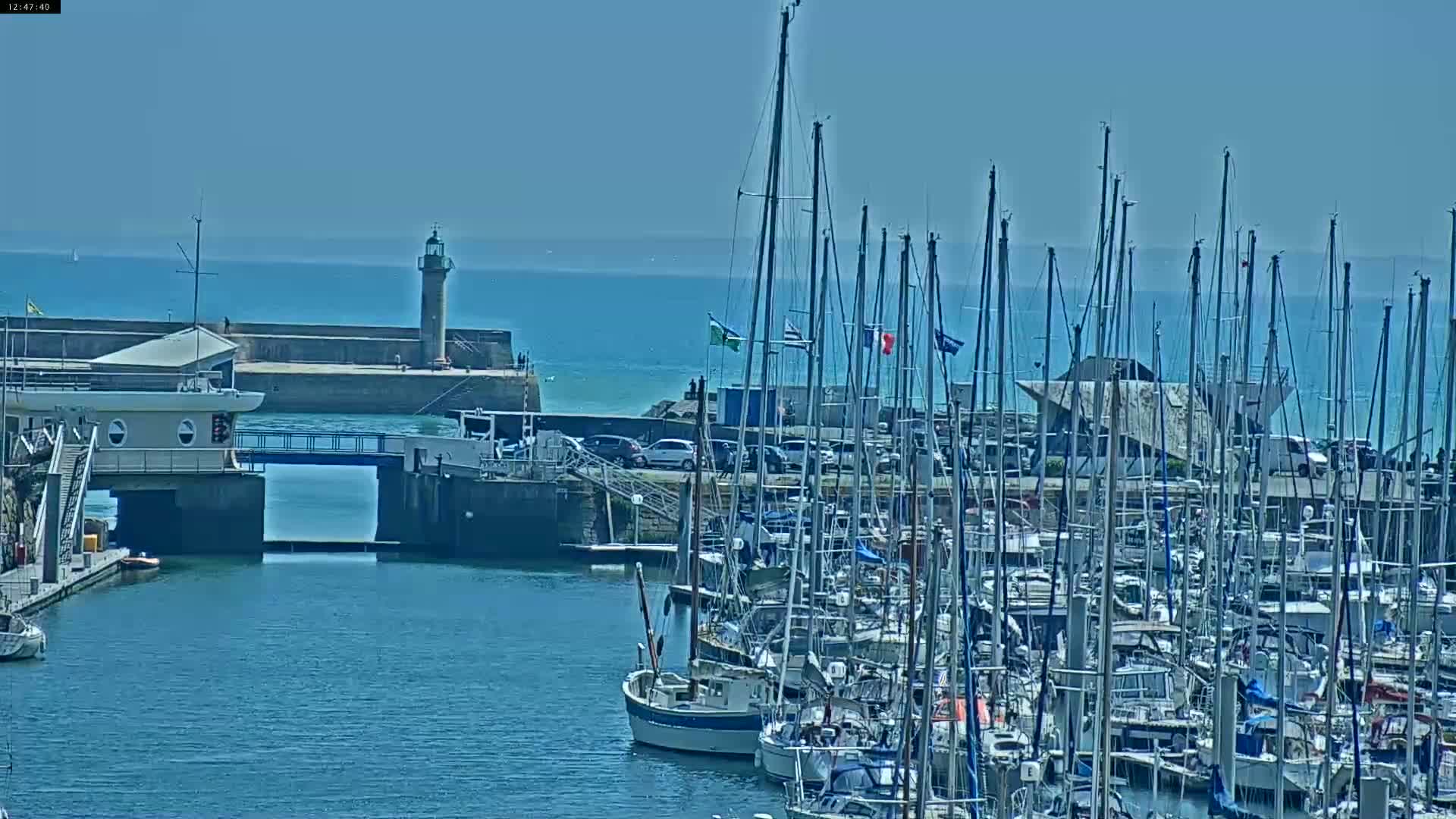 A harbor filled with numerous sailboats under a clear, sunny sky, features a lighthouse and a breakwater in the background.