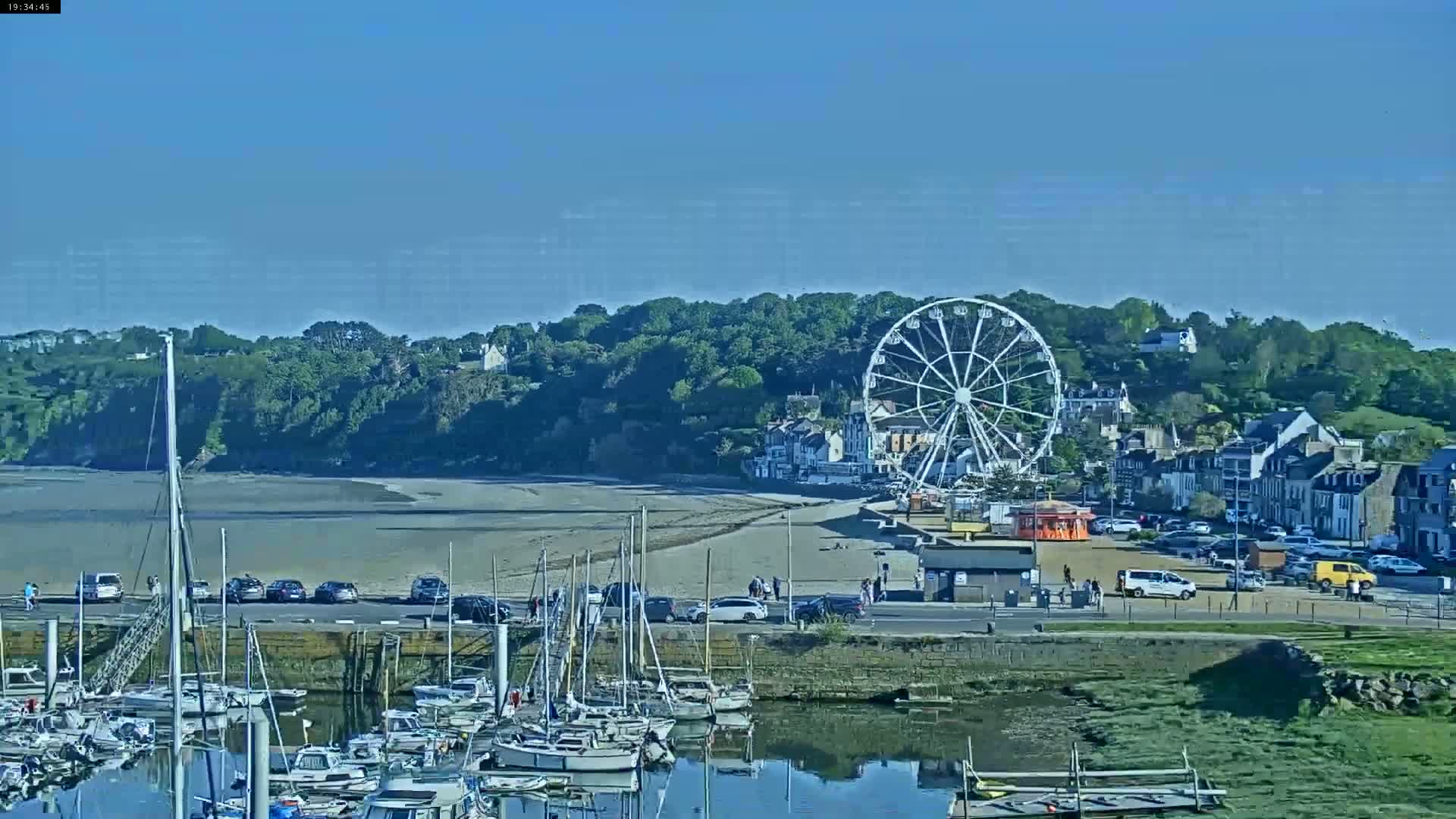 A Ferris wheel sits on a beach next to a marina filled with sailboats, under a clear blue sky.