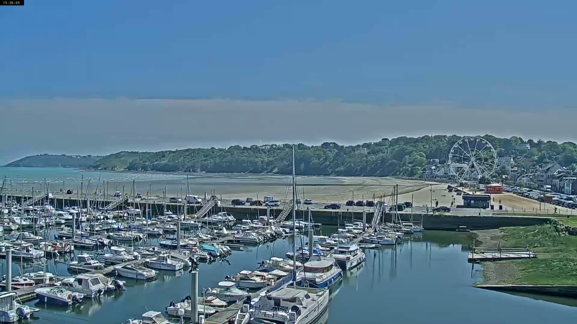 A marina filled with numerous boats is situated beside a sandy beach and a Ferris wheel under a clear, sunny sky.