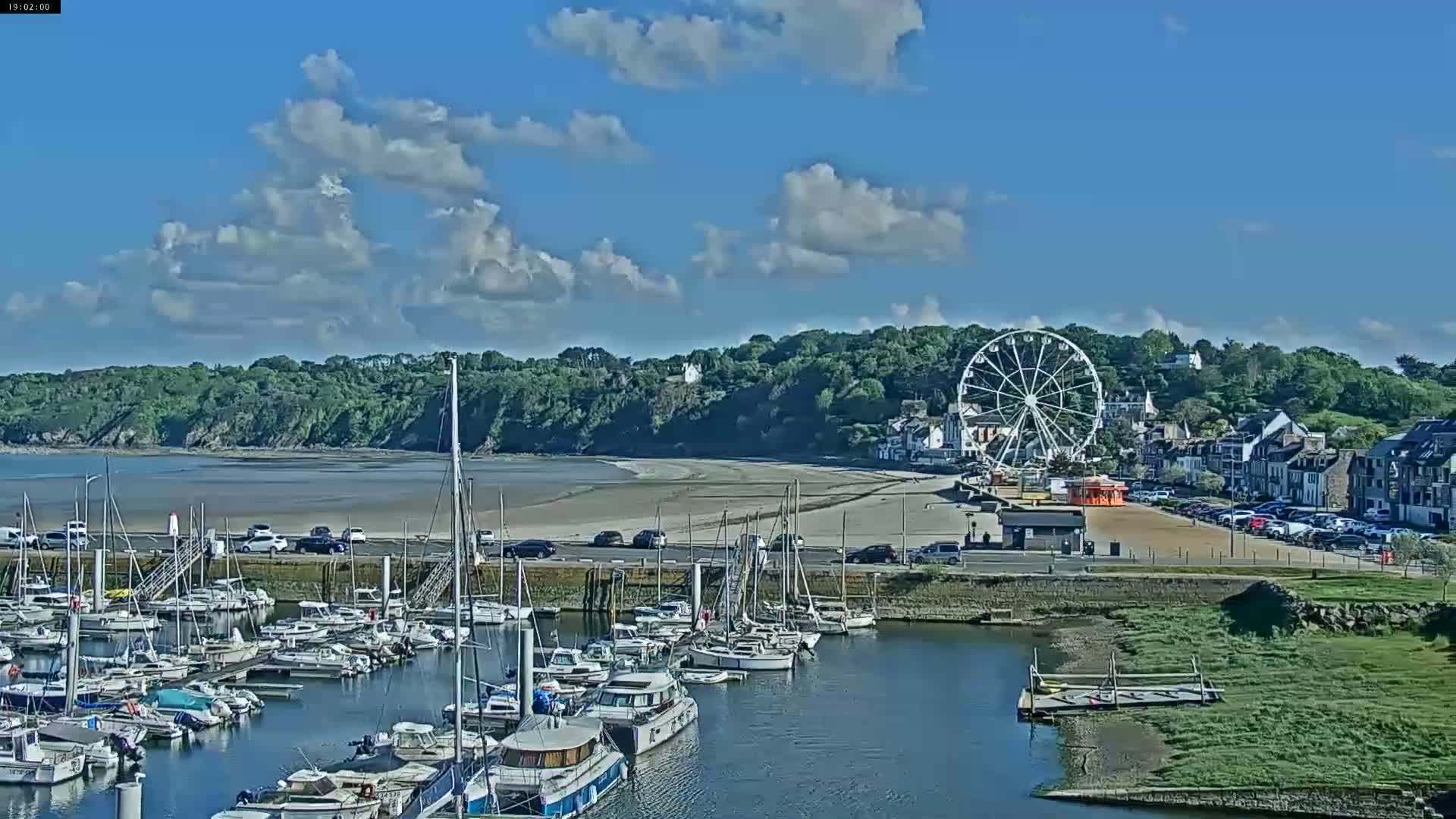 A marina filled with sailboats and motorboats is situated near a sandy beach, backed by a hillside with a Ferris wheel and buildings, under a mostly sunny sky with scattered clouds.
