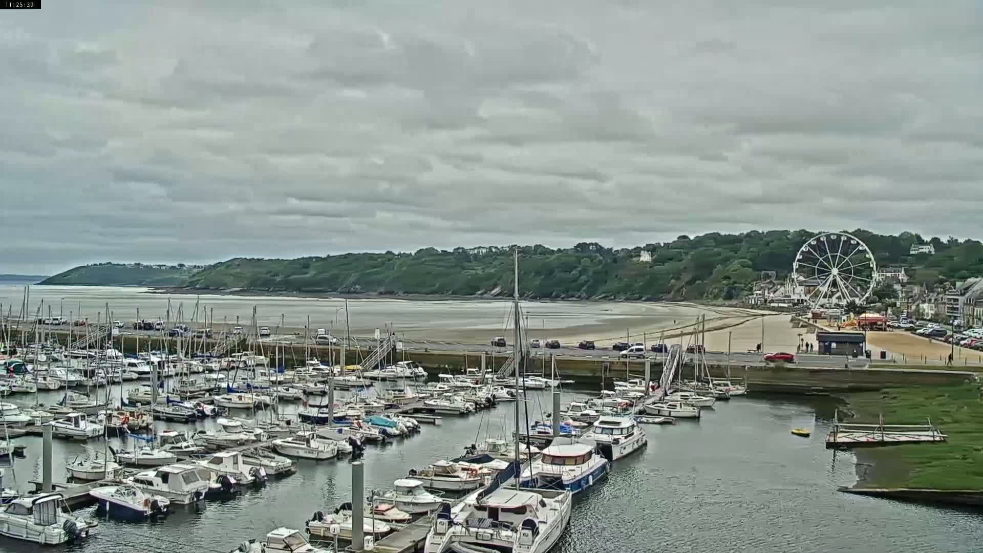 A marina filled with numerous boats is situated beside a sandy beach and a Ferris wheel under a cloudy sky.