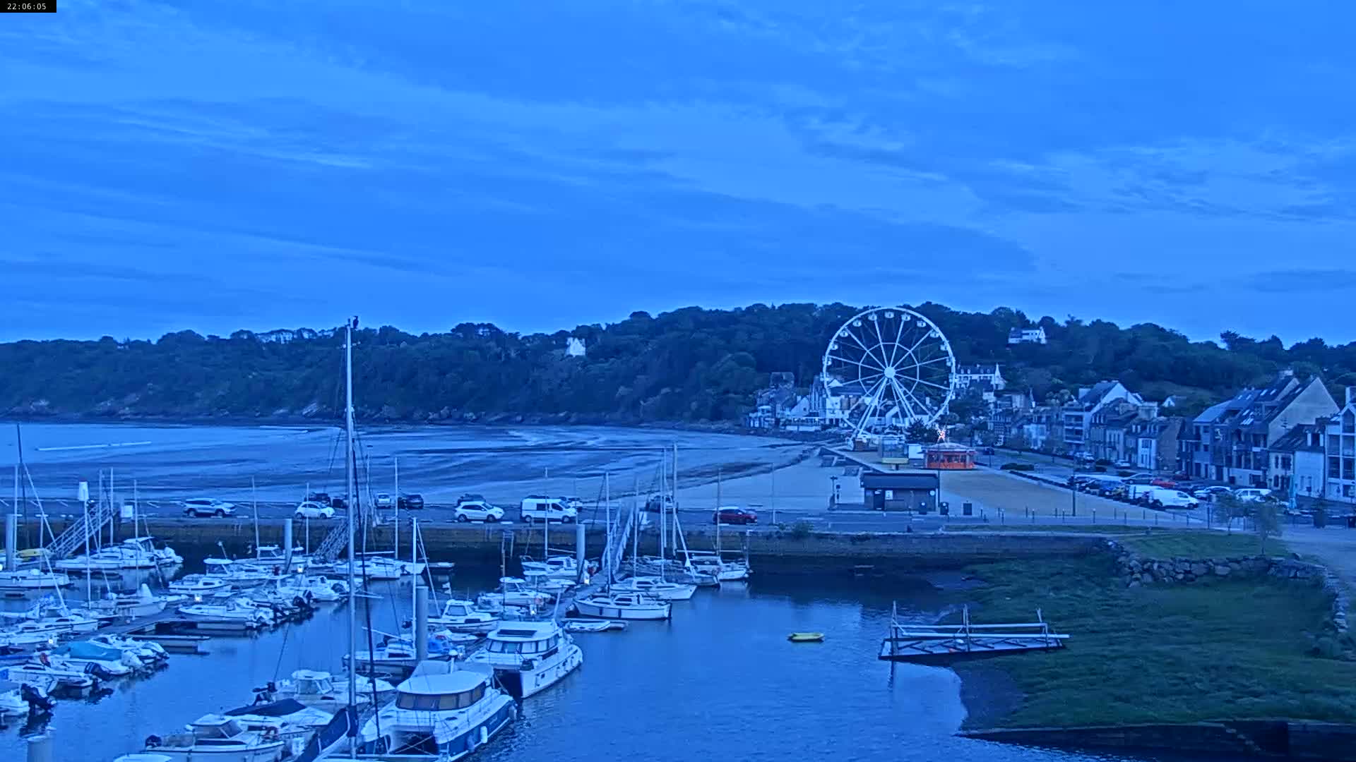 A marina filled with sailboats sits at the base of a hill with a Ferris wheel and buildings under a twilight blue sky.