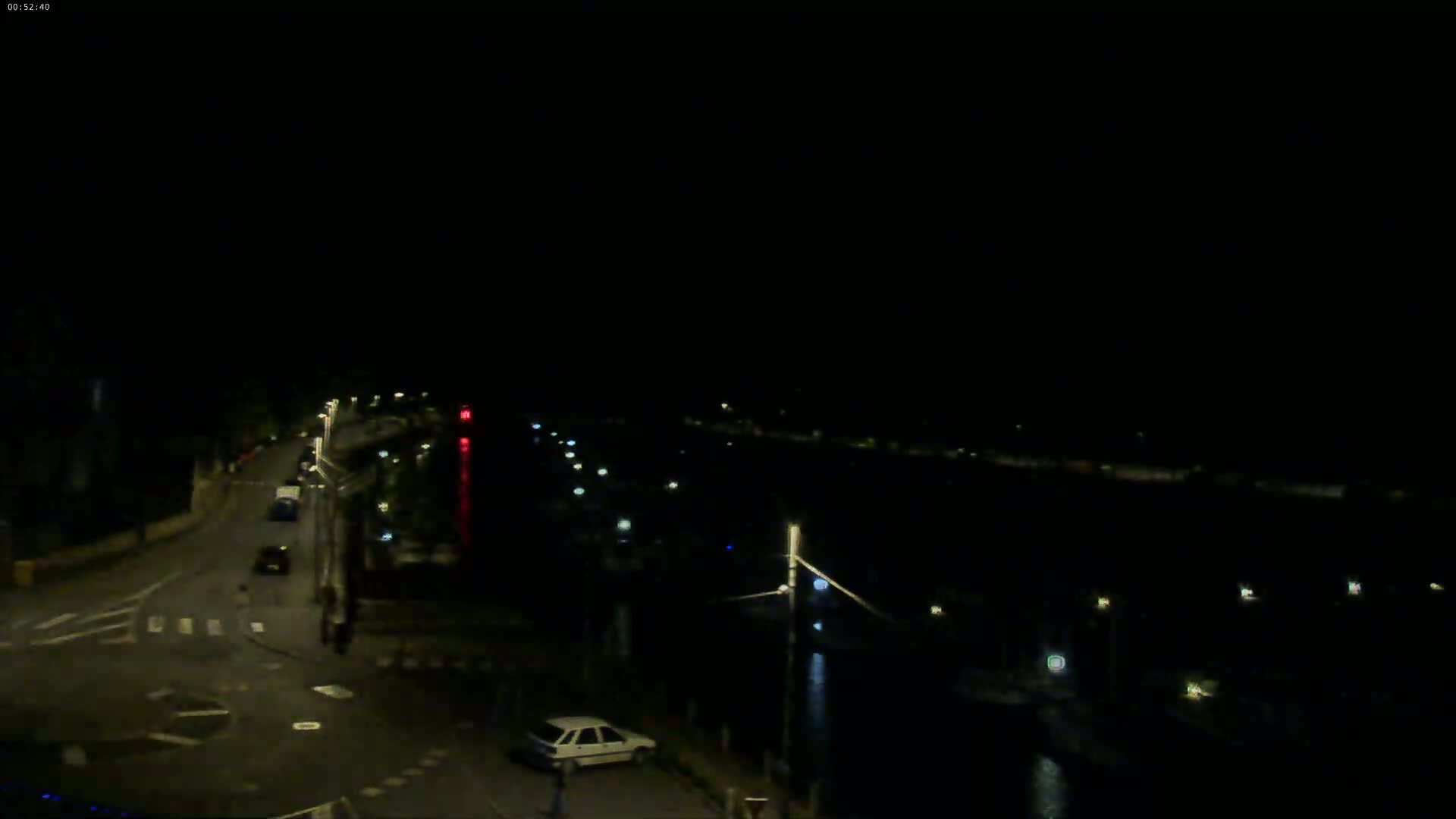 A nighttime, high-angle view shows a road with a few cars, intersecting with another road near a dark body of water with sparsely lit boats along its edge.