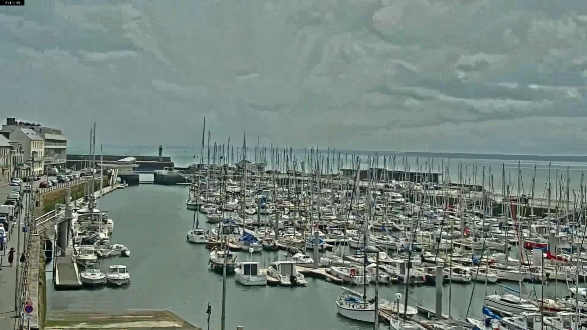 A marina filled with numerous sailboats and motorboats is situated on a calm body of water under a mostly cloudy sky.
