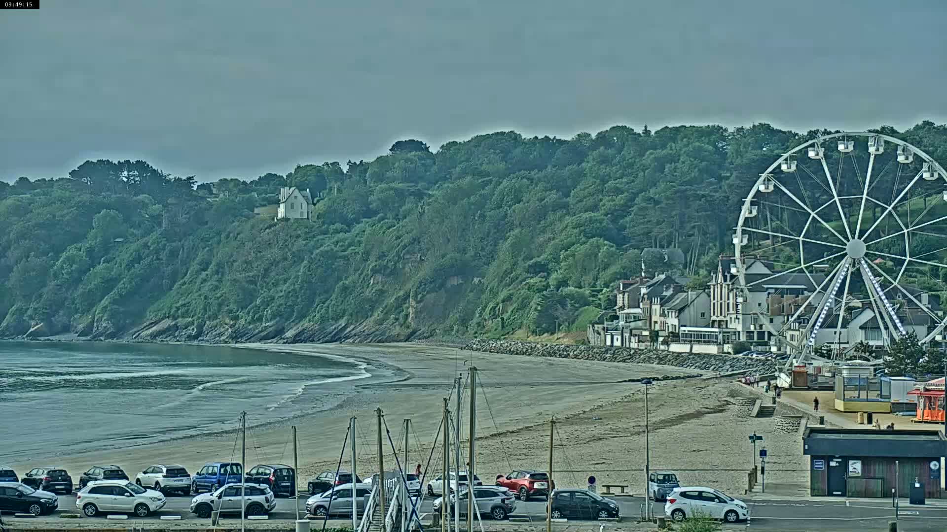 A sandy beach with calm water, parked cars, and a Ferris wheel sits before a lush green hillside under an overcast sky.