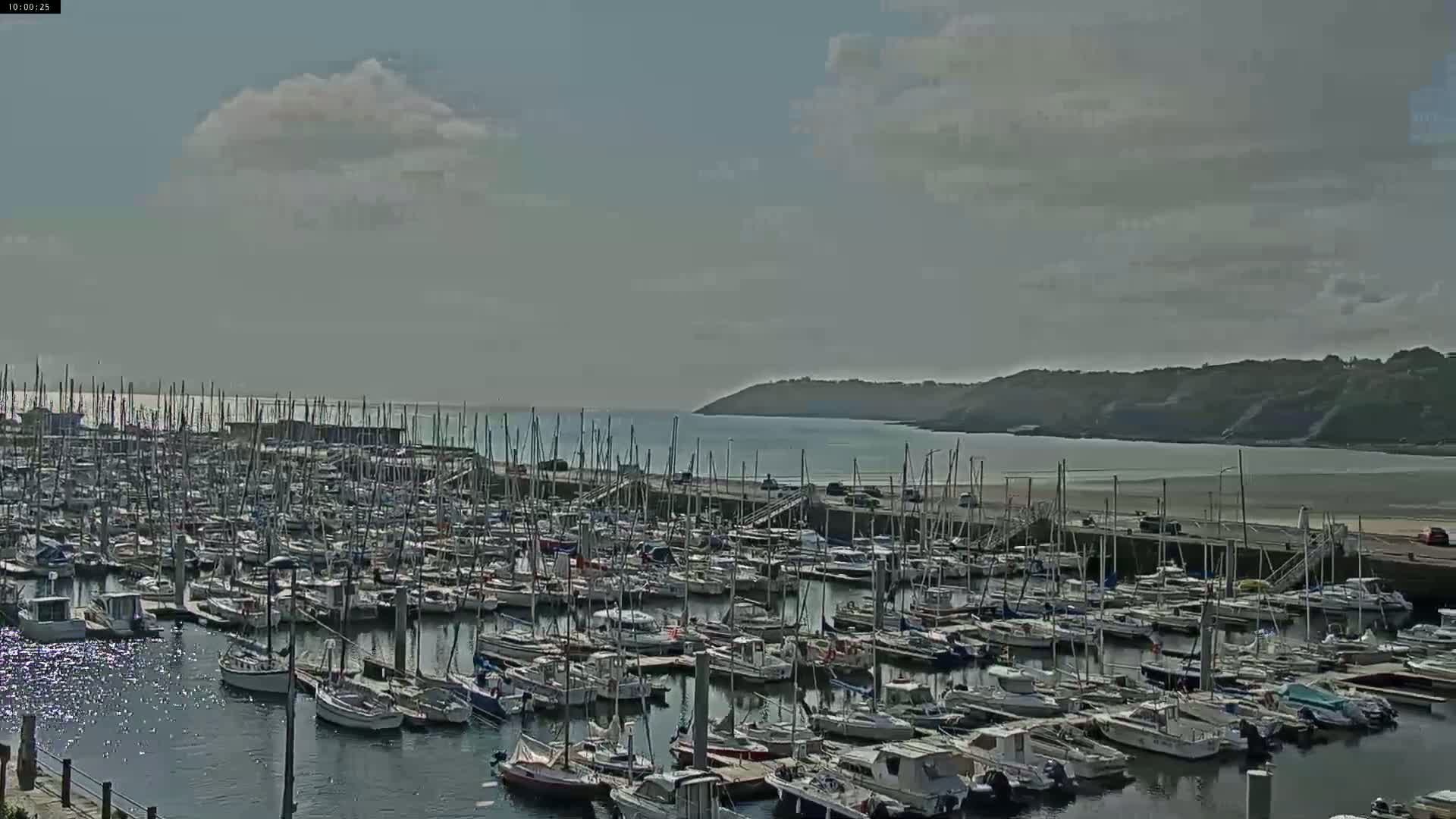 A large marina filled with many sailboats is situated by a calm body of water under a partly cloudy sky.