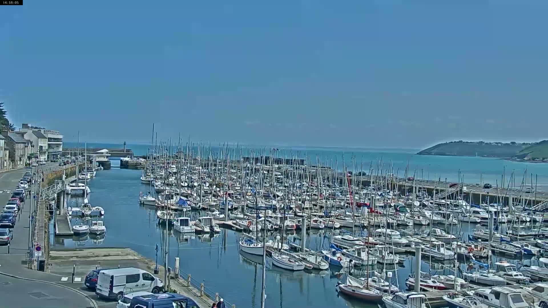 A marina filled with numerous sailboats and motorboats is seen on a sunny day, with calm waters and a distant shoreline visible.