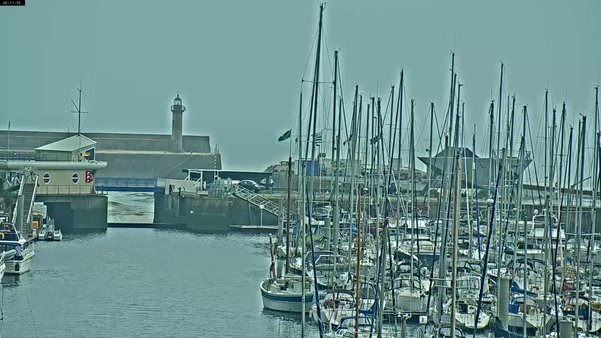 A harbor with many sailboats moored, a lighthouse in the distance, and overcast skies.