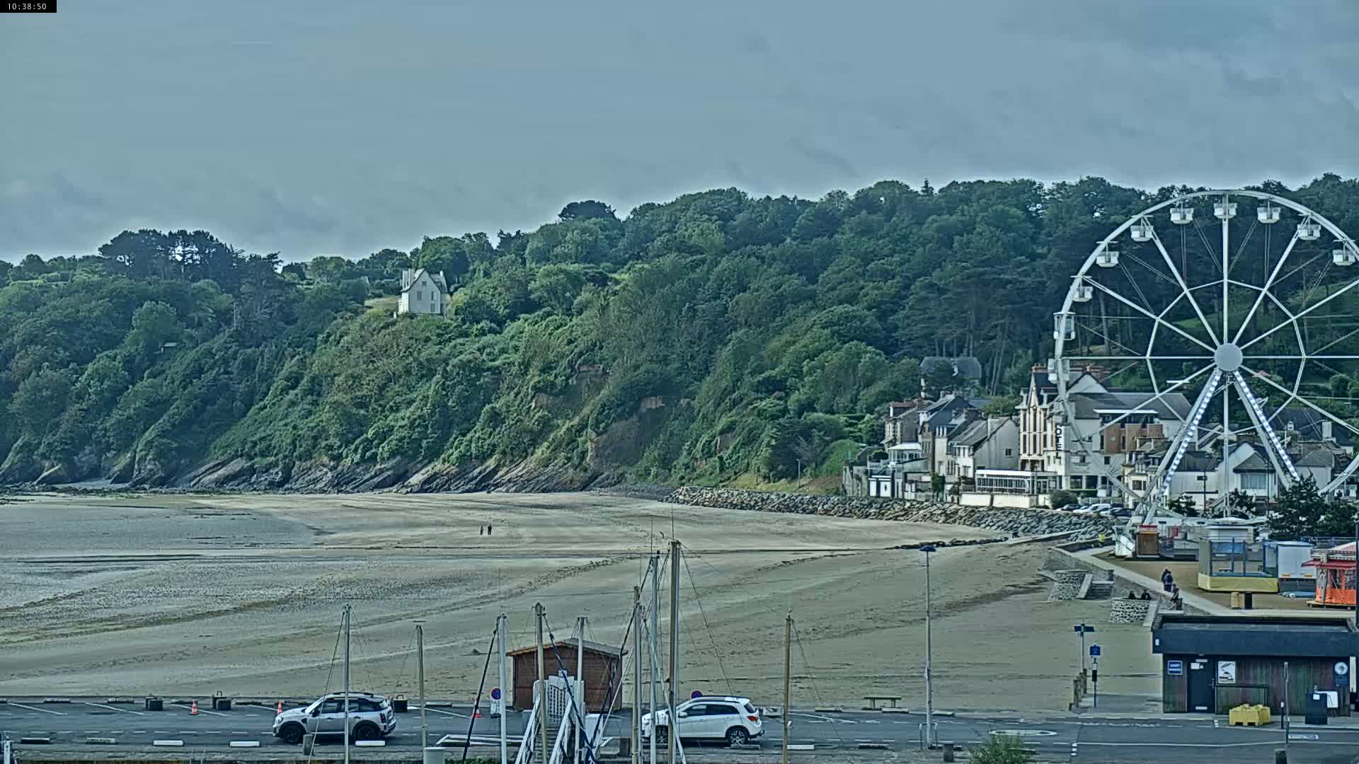 A sandy beach stretches between a green hillside with buildings and a Ferris wheel on the right, under an overcast sky.
