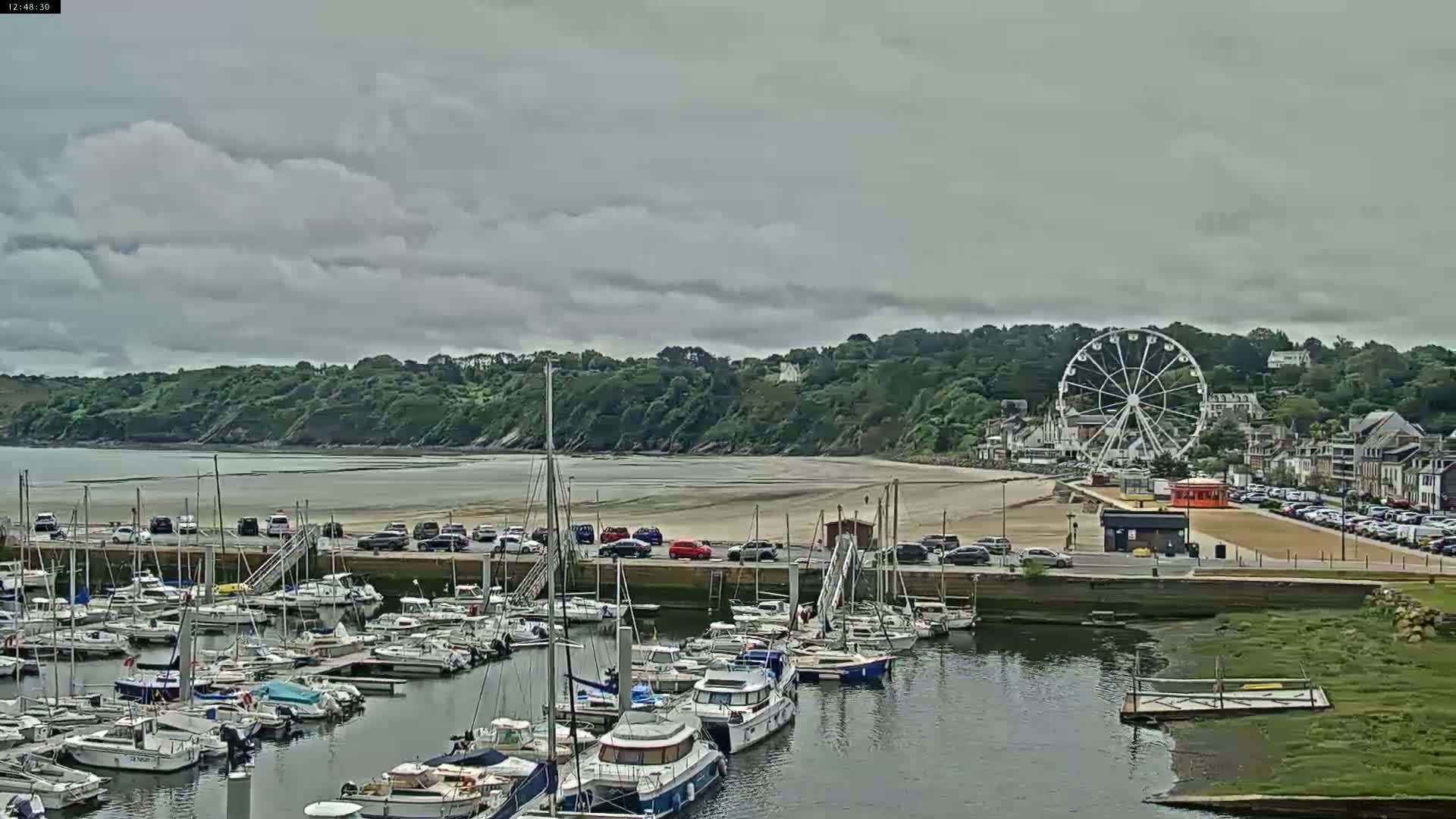 A harbor filled with numerous boats sits beside a sandy beach and a Ferris wheel under a cloudy sky.