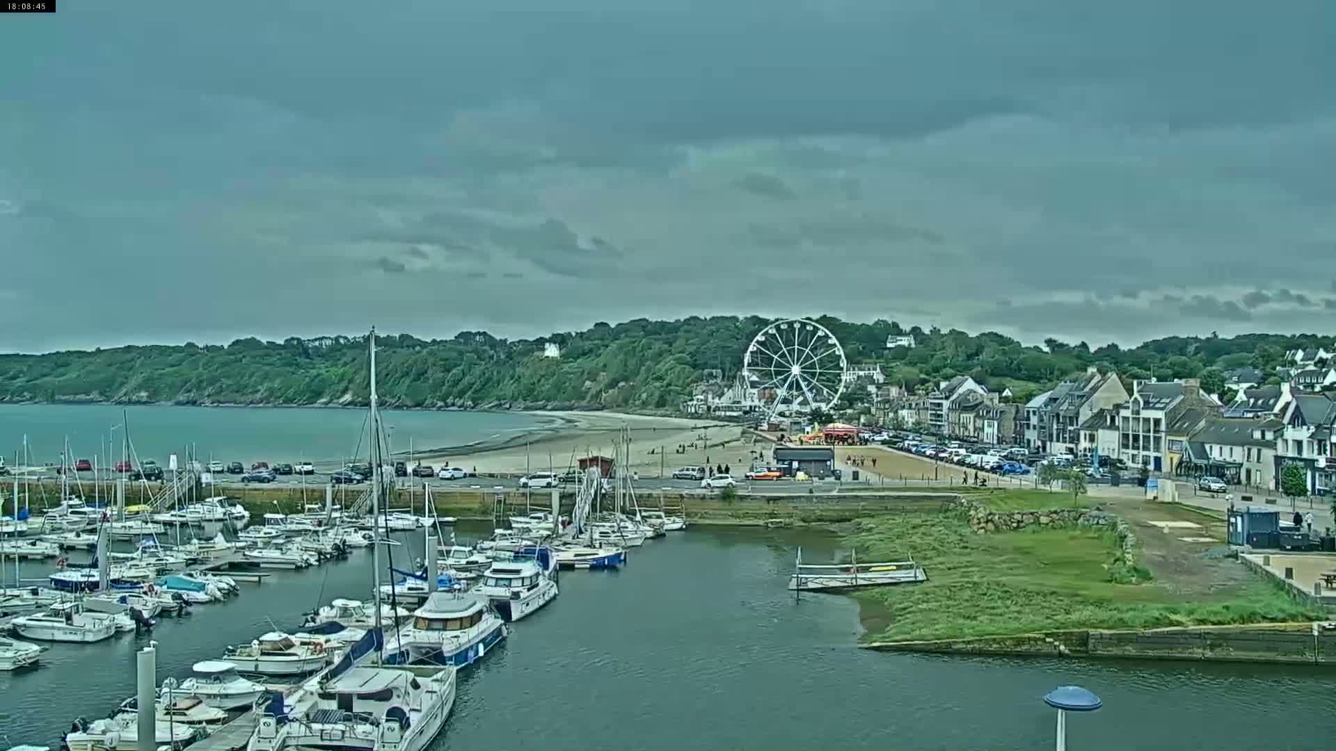 A marina filled with sailboats and motorboats is situated next to a beach with a Ferris wheel and seaside town under an overcast sky.