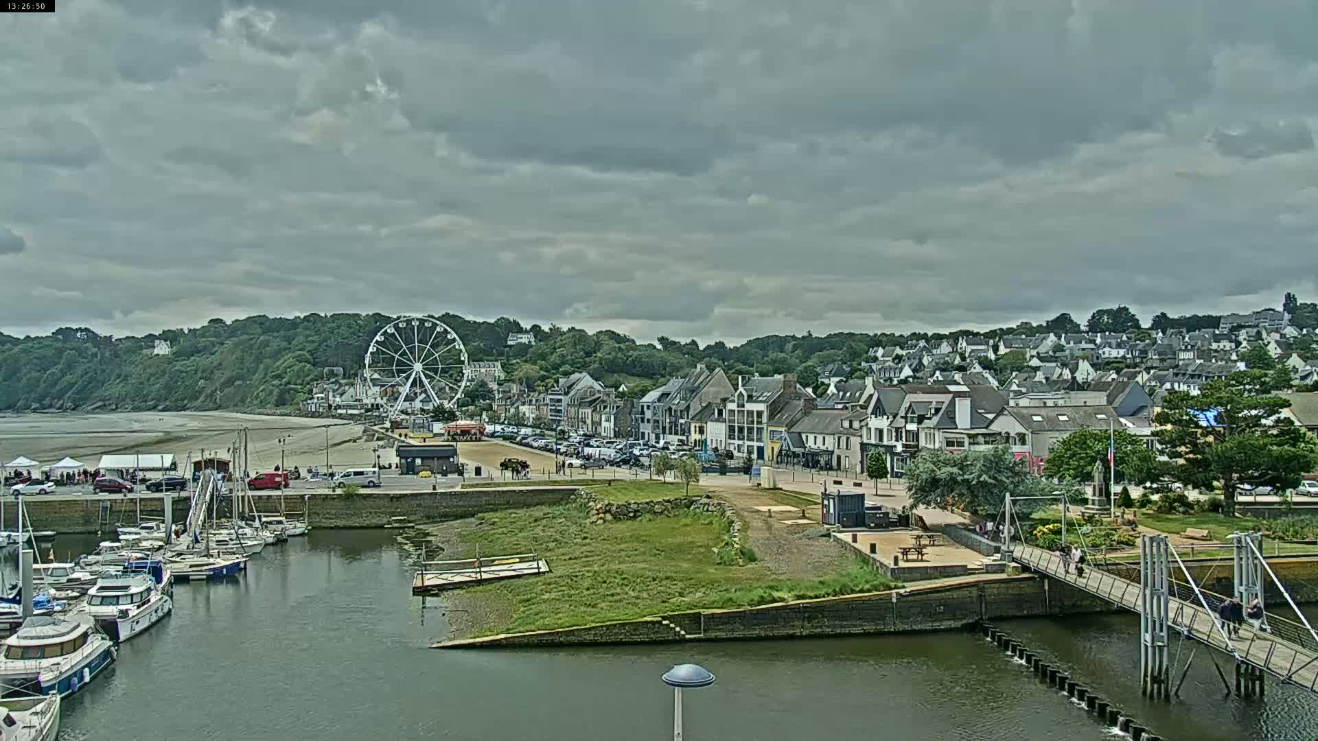 A harbor filled with boats sits below a coastal town under a cloudy sky, with a Ferris wheel visible in the distance.