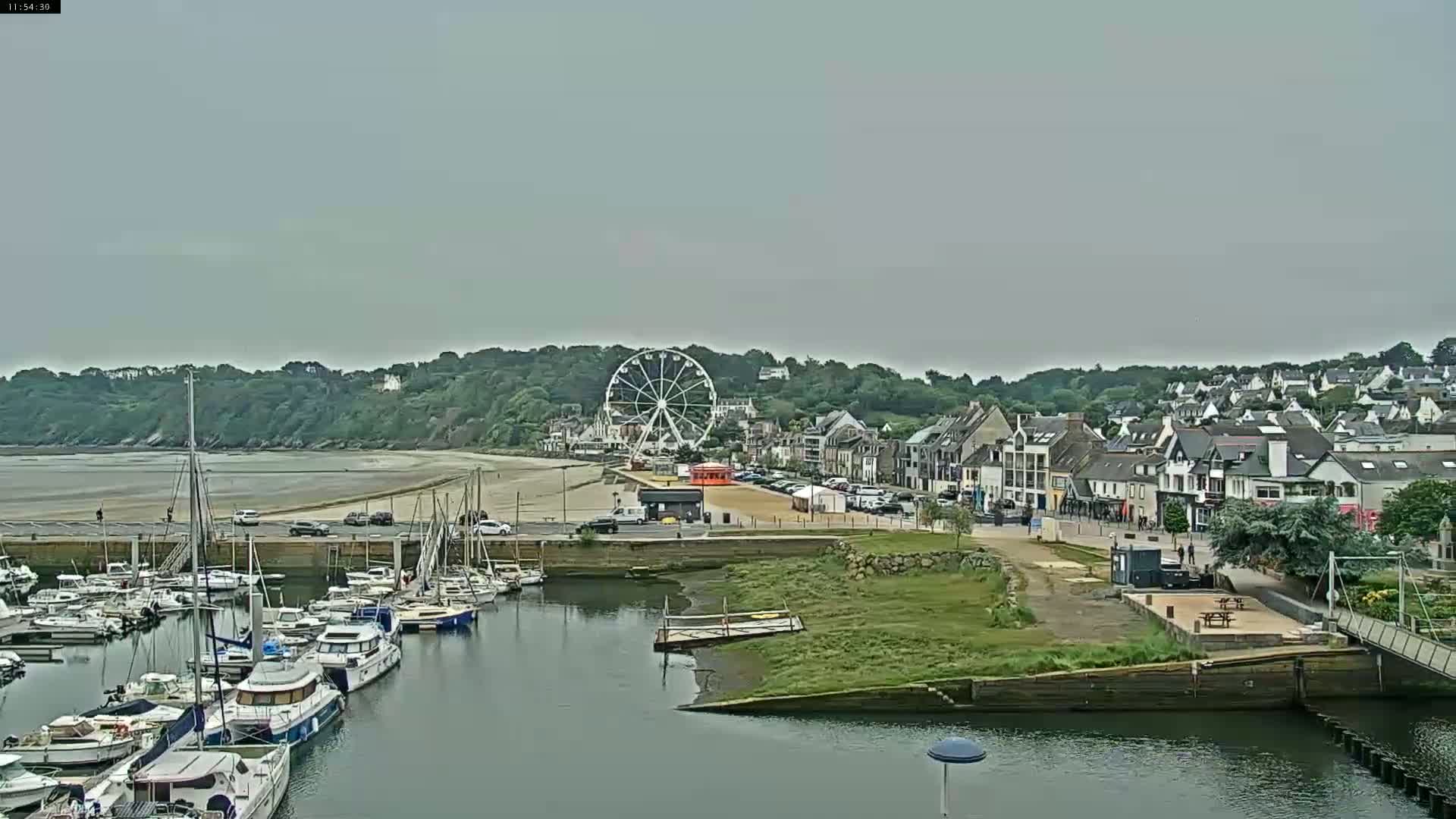 A harbor filled with sailboats and motorboats is situated near a town with a Ferris wheel on a beach under an overcast sky.