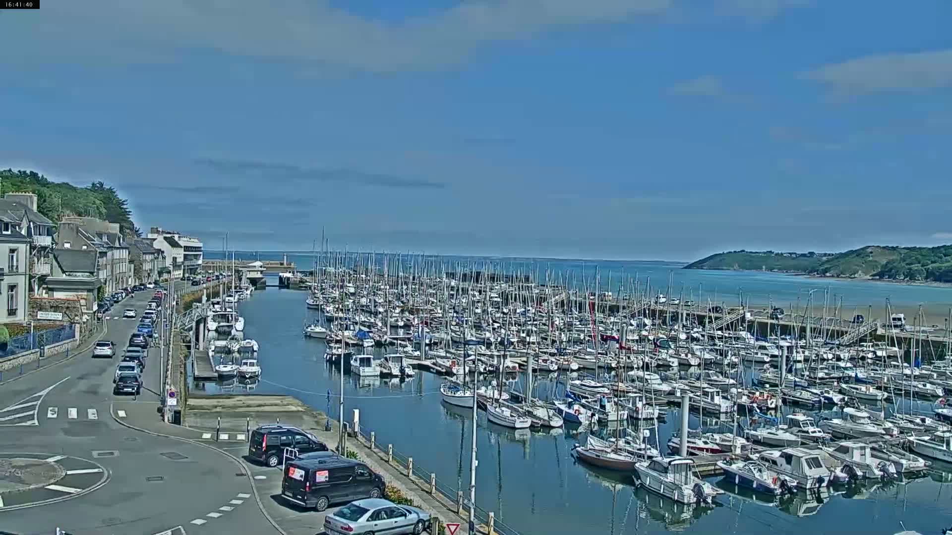 A sunny day shows a harbor filled with numerous sailboats and motorboats, a road with cars lining the harbor's edge, and buildings along the waterfront.