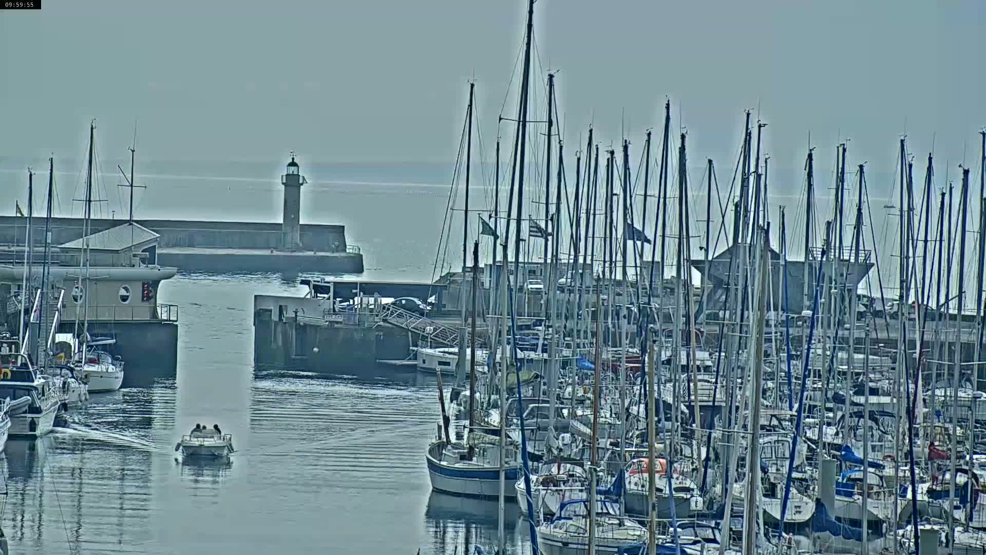 A harbor filled with numerous sailboats under an overcast sky, with a lighthouse visible in the distance.