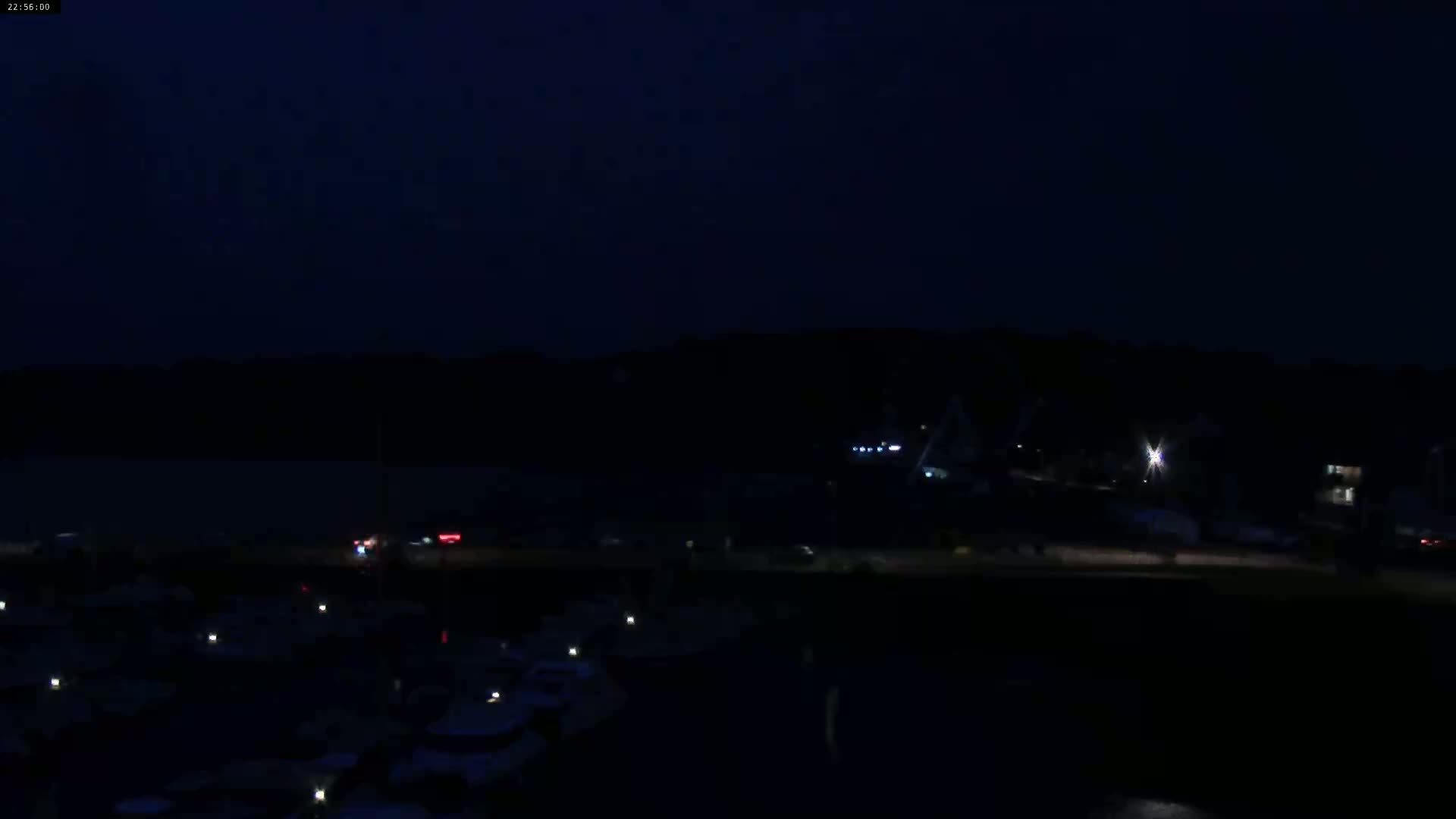 A dark nighttime scene shows a body of water with docked boats, a shoreline with sparse lights, and a hill in the background under a mostly clear, dark sky.