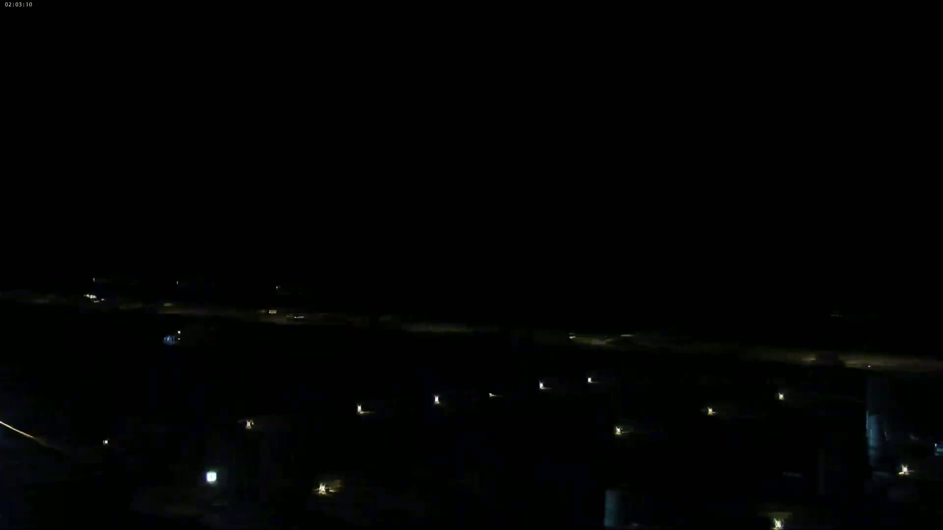 A dark nighttime scene shows a body of water with docked boats, a shoreline with sparse lights, and a hill in the background under a mostly clear, dark sky.