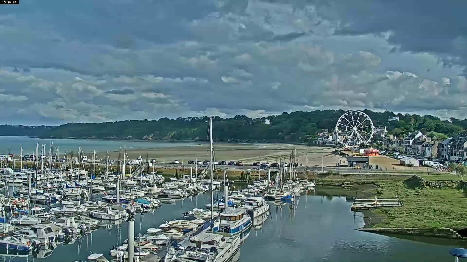 A marina filled with numerous boats is situated near a beach with a Ferris wheel under a partly cloudy sky.
