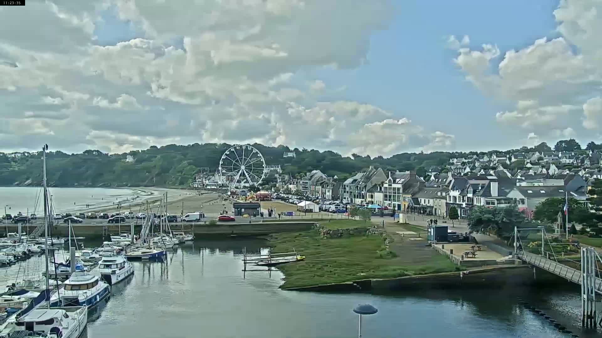 A seaside town with a Ferris wheel is visible on a partly cloudy day, with a marina filled with boats in the foreground and a sandy beach in the background.