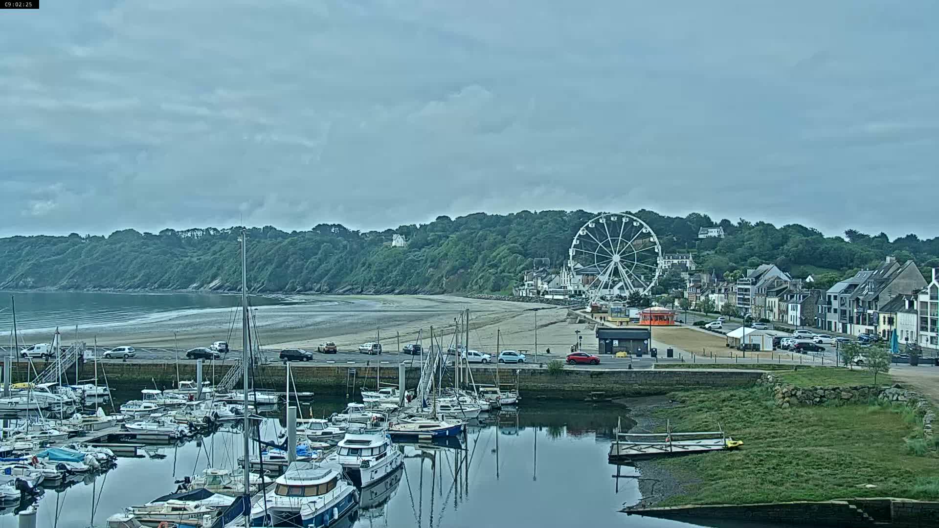 A marina filled with sailboats and motorboats is situated next to a sandy beach, backed by a hill with buildings and a Ferris wheel, under a cloudy sky.