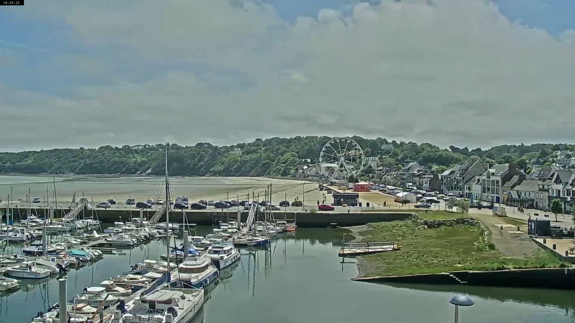 A marina filled with sailboats and motorboats is situated next to a sandy beach, beyond which is a town with a Ferris wheel under a partly cloudy sky.