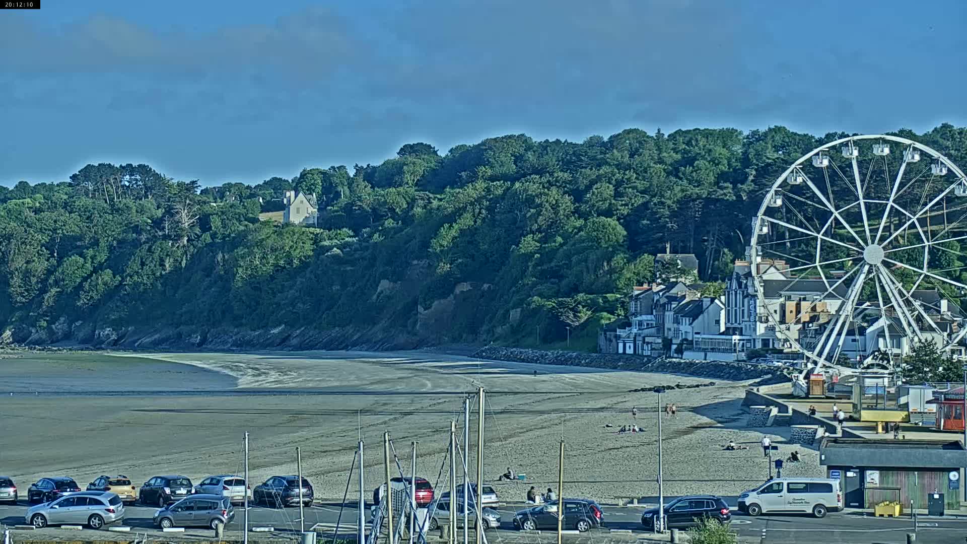 A mostly sunny day reveals a beach with parked cars, a Ferris wheel, and a few people relaxing, all situated below a lush green cliffside.
