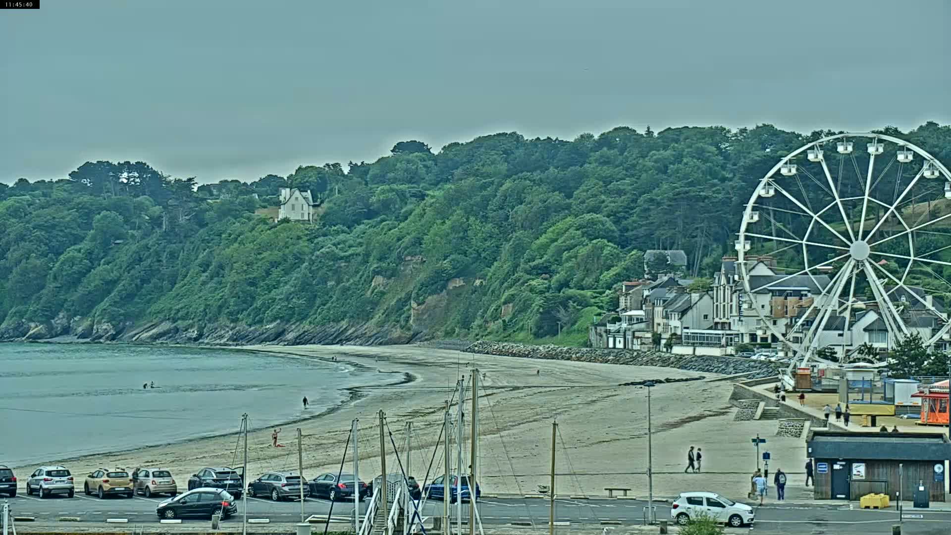 A sandy beach stretches along a calm bay, backed by a green cliff with buildings and a Ferris wheel, under an overcast sky.