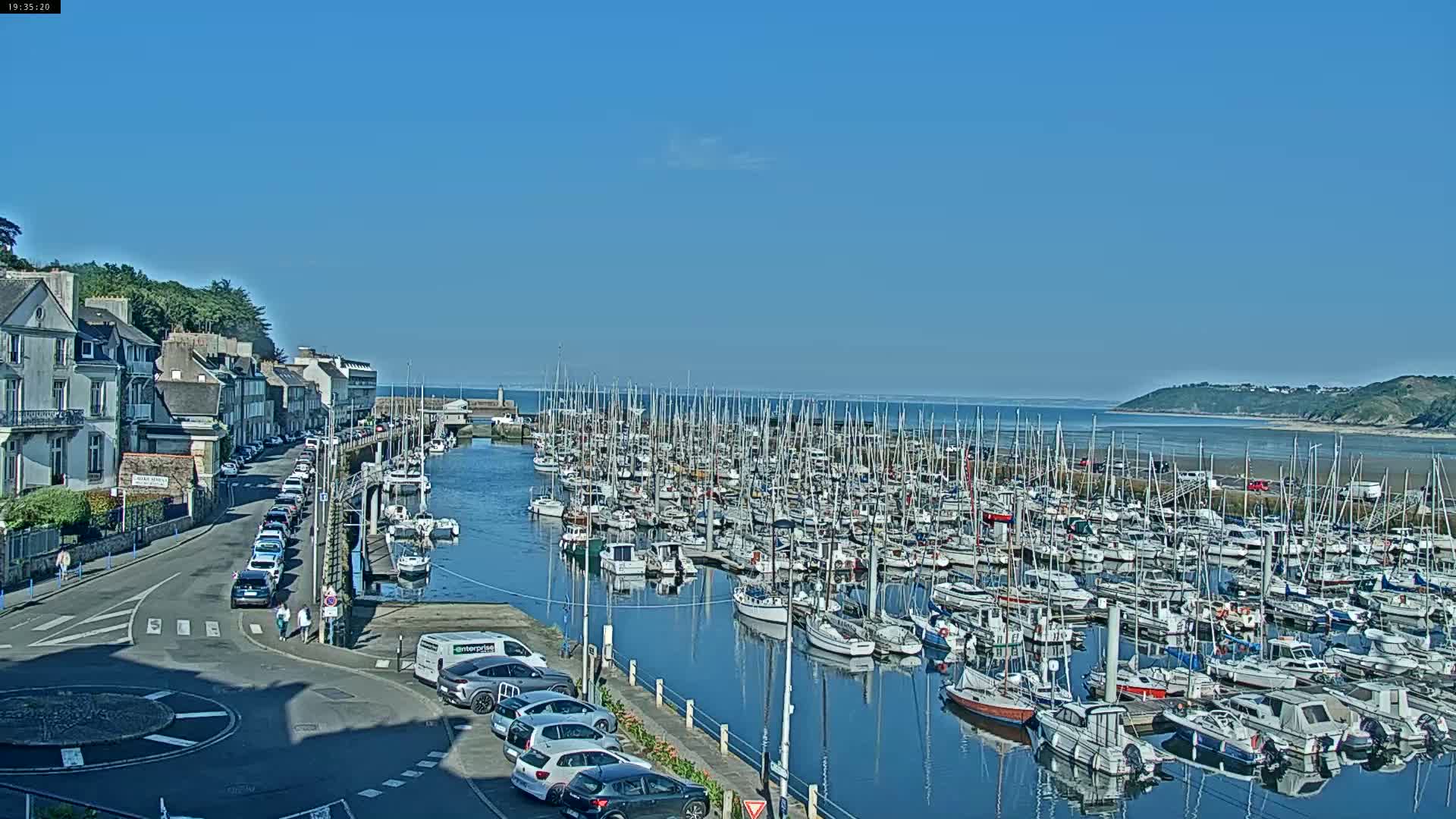 A harbor filled with numerous sailboats and yachts is situated beside a town with buildings lining a street, all under a clear, sunny sky.