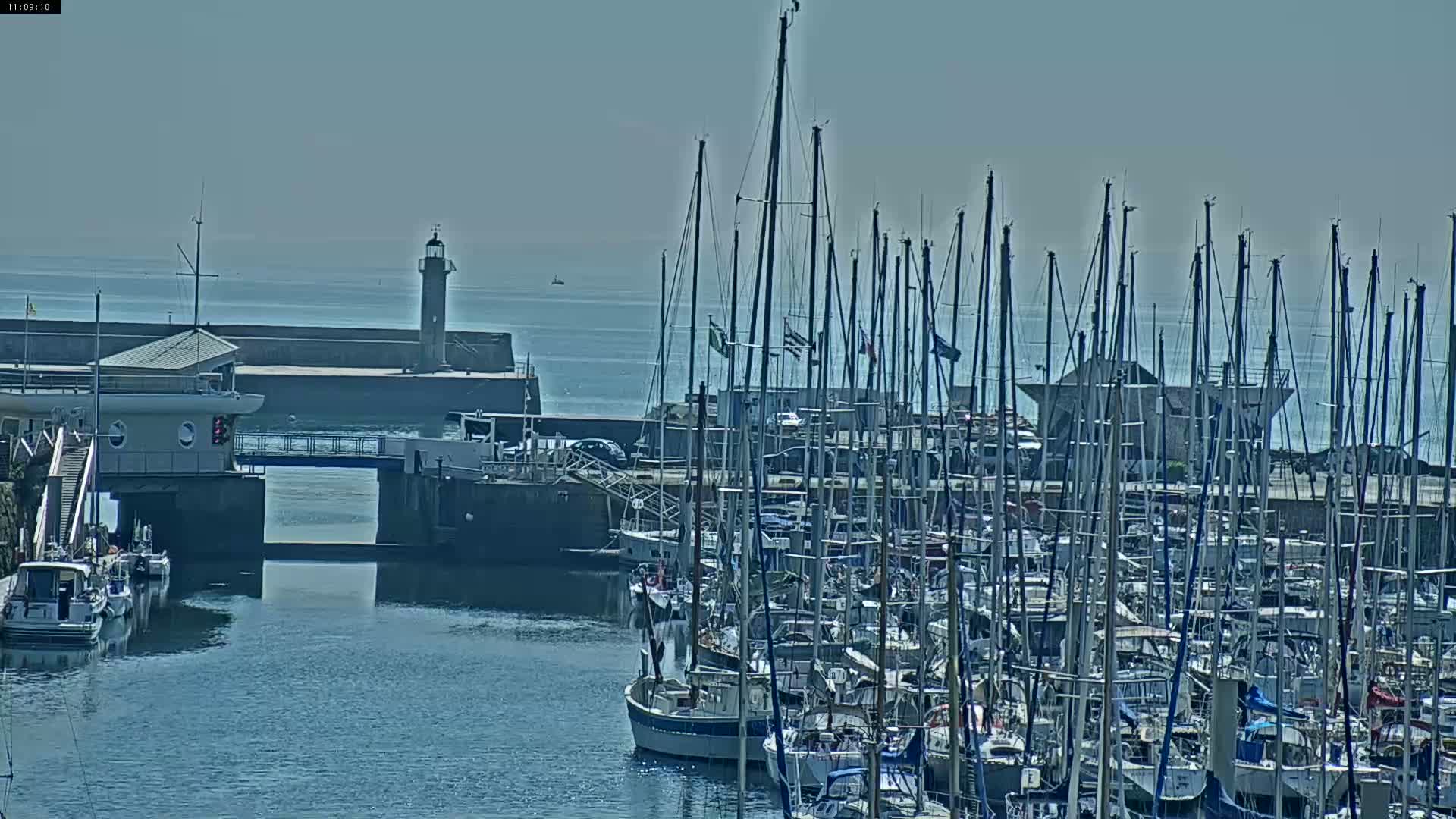 Many sailboats are docked in a harbor on a sunny day, with a lighthouse visible in the distance.