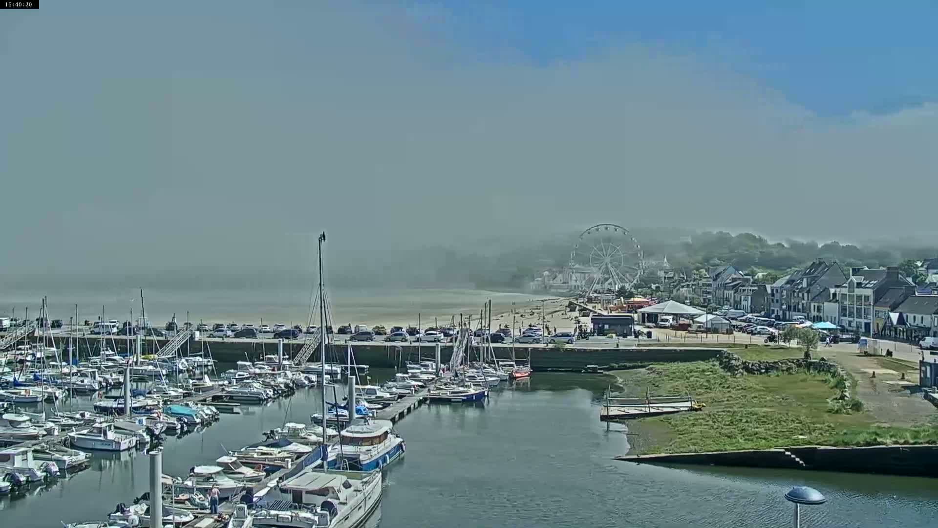 A hazy day reveals a marina filled with sailboats and motorboats, a beach with people, a Ferris wheel, and buildings along a shoreline.