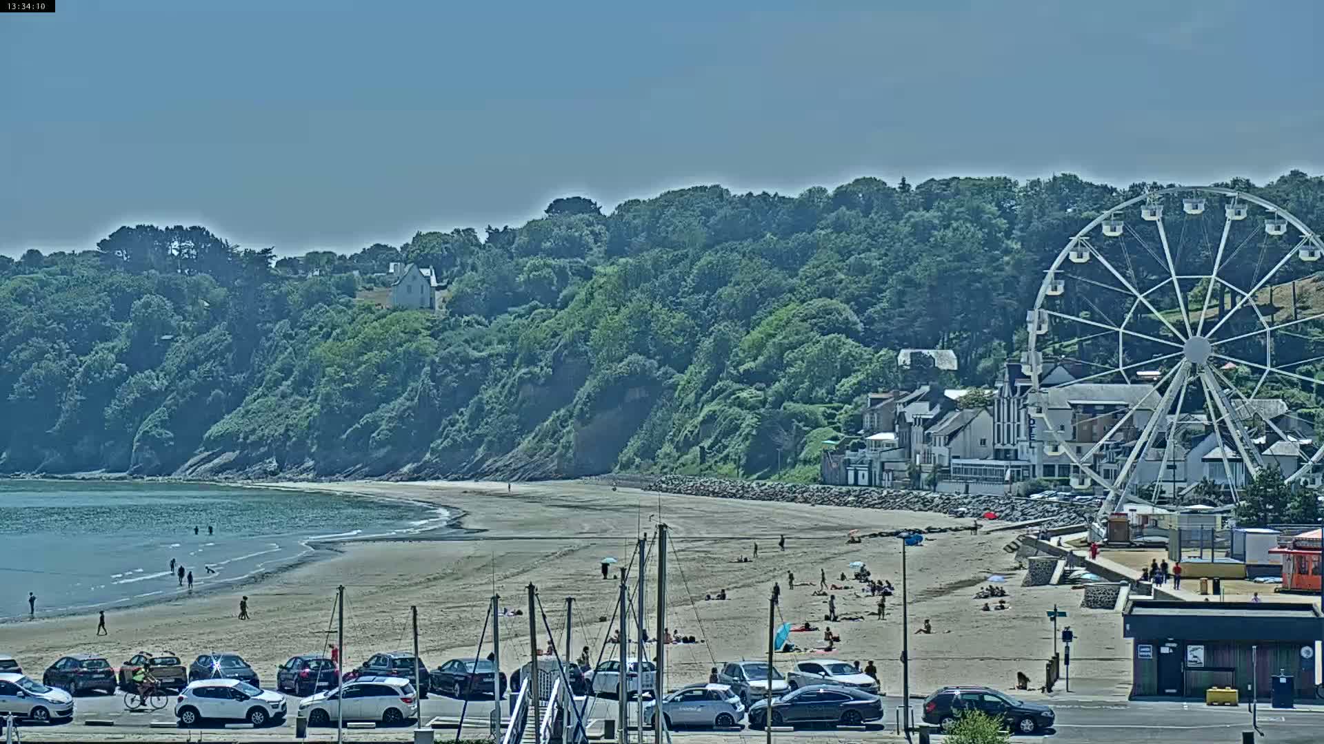 A sandy beach with people sunbathing and swimming, backed by a grassy hillside with buildings and a Ferris wheel, under a clear blue sky.