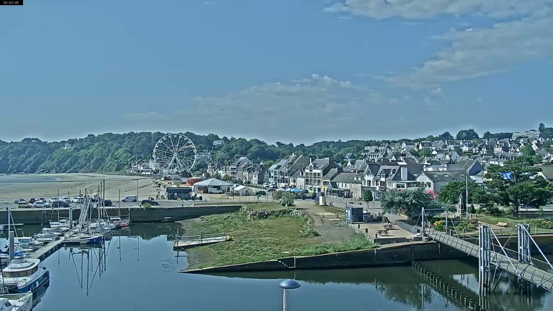 A seaside town on a partly sunny day features a marina with boats, a sandy beach, a Ferris wheel, and numerous buildings, all nestled beside a verdant hill.