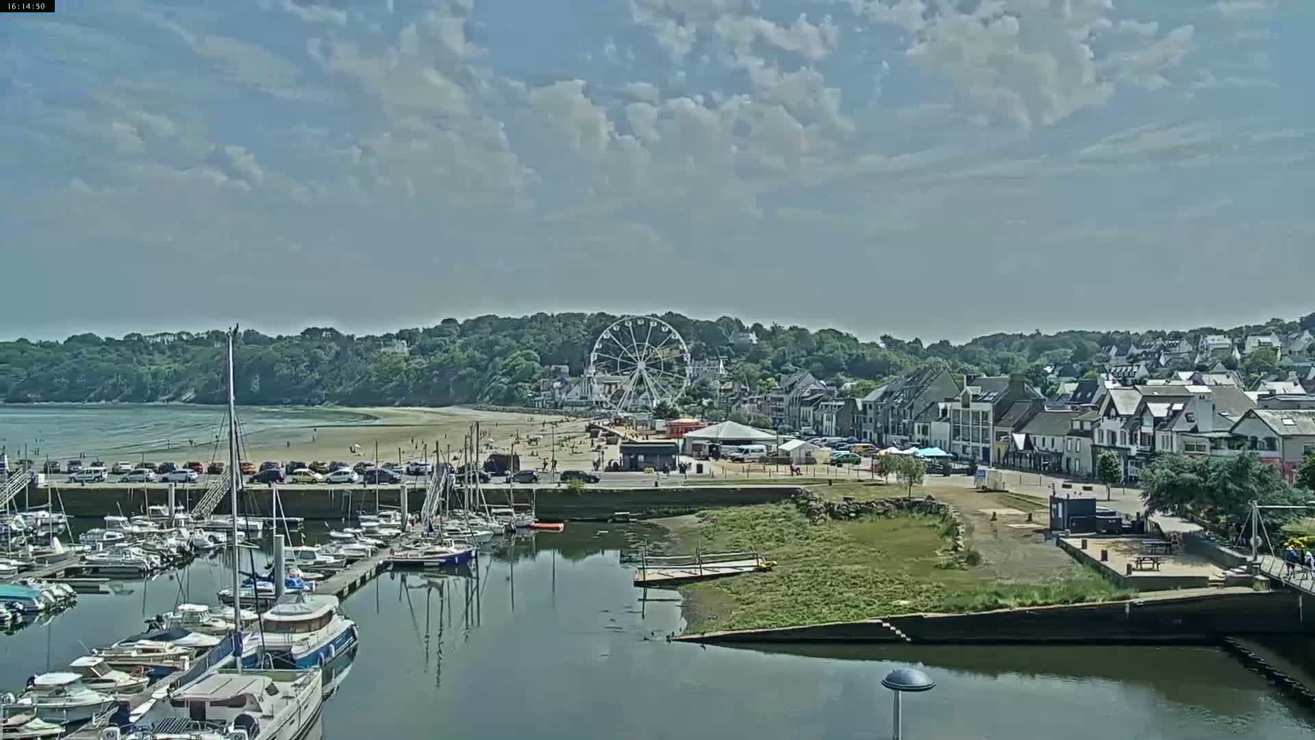 A harbor filled with sailboats is situated near a sandy beach and a town with a Ferris wheel, under a partly cloudy sky.