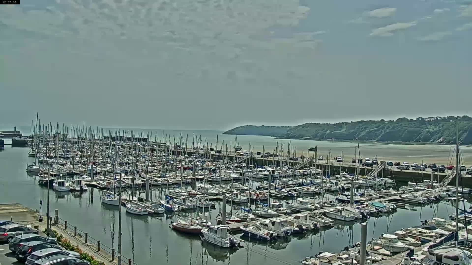 A marina filled with numerous sailboats and motorboats is situated on calm water under a partly cloudy sky.
