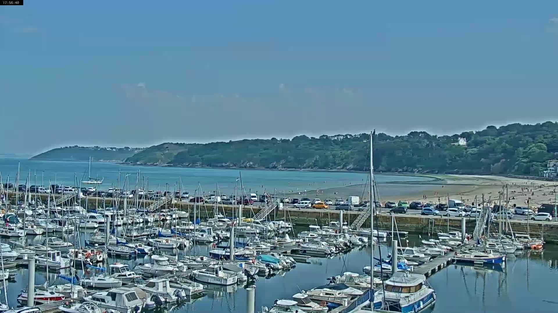A marina filled with numerous boats is situated near a sandy beach and a calm bay under a clear, sunny sky.