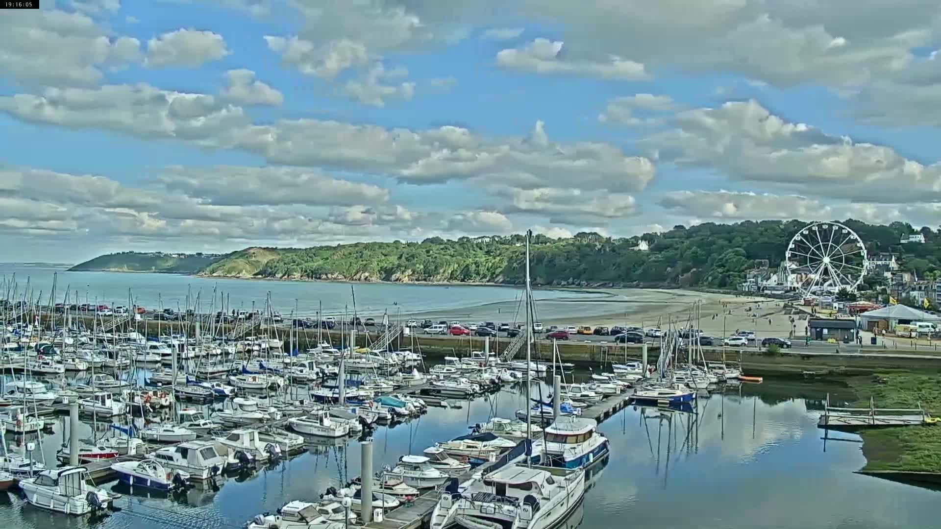 A marina filled with sailboats and motorboats is situated next to a beach with a Ferris wheel, under a partly cloudy sky.