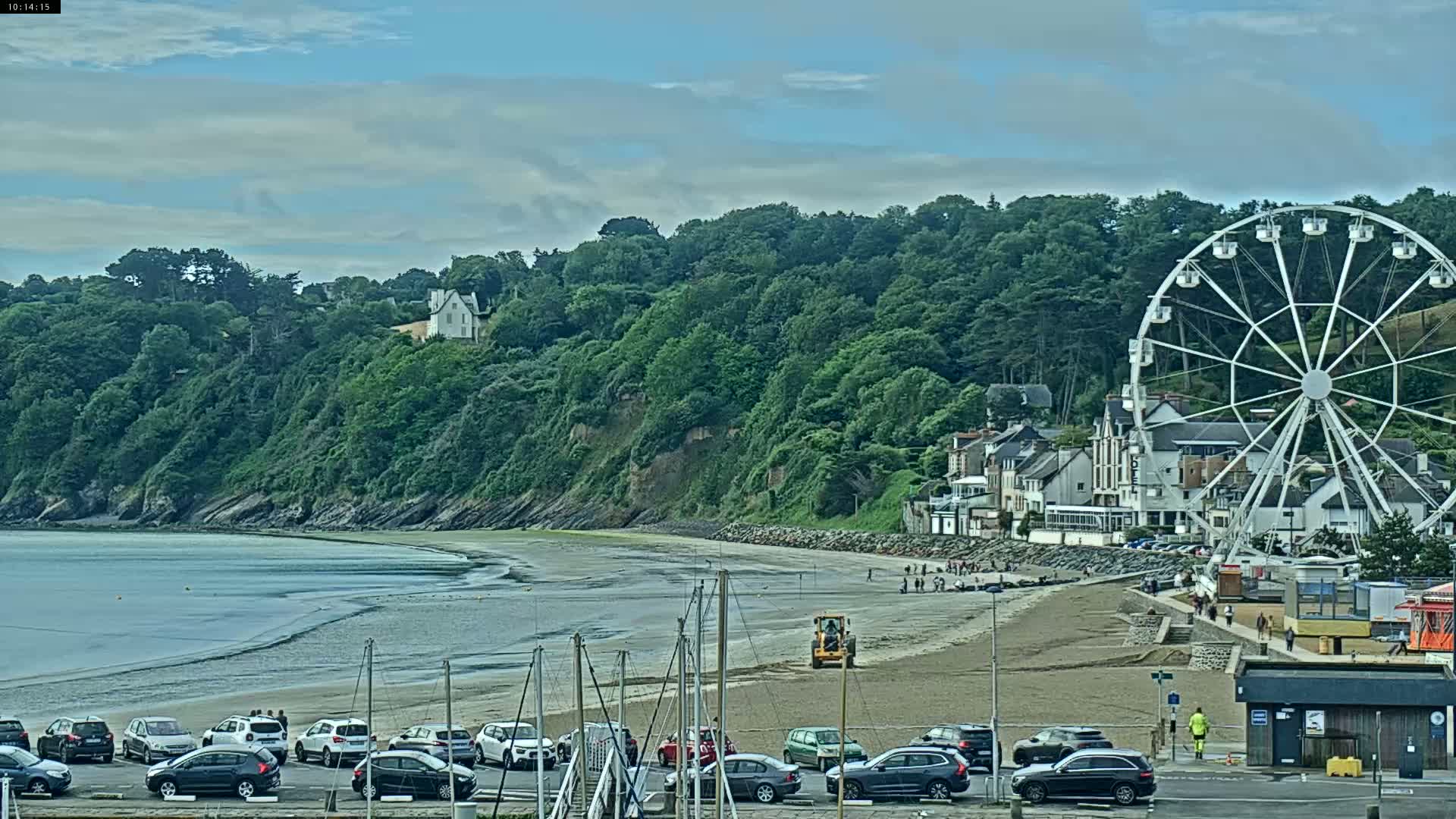 A beach with a Ferris wheel and cars parked along the shore, backed by a lush green hillside under a partly cloudy sky.