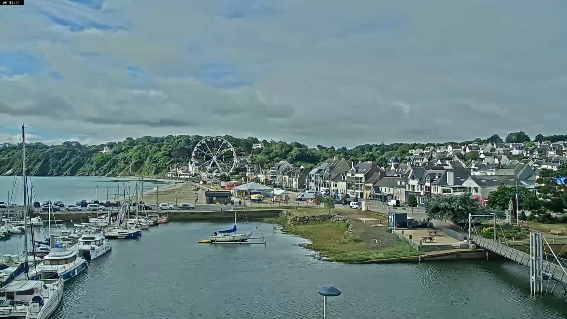 A harbor filled with sailboats is situated next to a town with a Ferris wheel, under a partly cloudy sky.