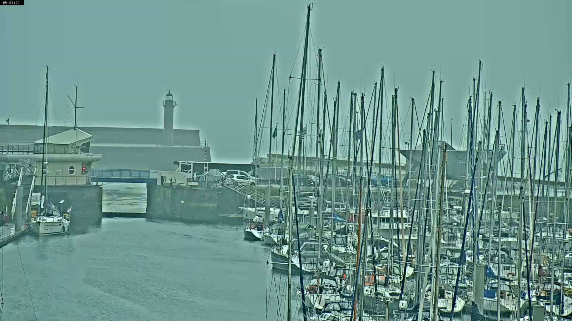 A marina filled with numerous sailboats under an overcast sky, with a lighthouse visible in the distance.